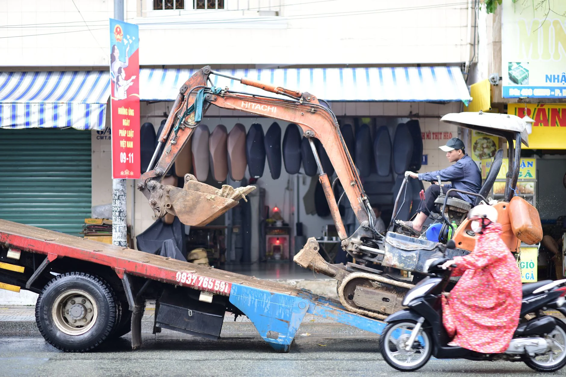 Excavator being loaded onto truck with motorbike passing by in urban street.